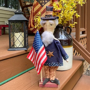 May include: A patriotic Uncle Sam doll stands on a porch, holding a small American flag. He wears a blue coat, striped pants, and a star-adorned hat. An American flag and yellow flowers are in the background.