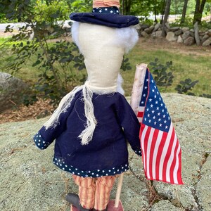 May include: A patriotic Uncle Sam doll, featuring a navy blue coat with white stars, striped pants, and a tall hat. The doll holds a small American flag. The doll is standing on a red wooden base. The doll has white hair and beard.