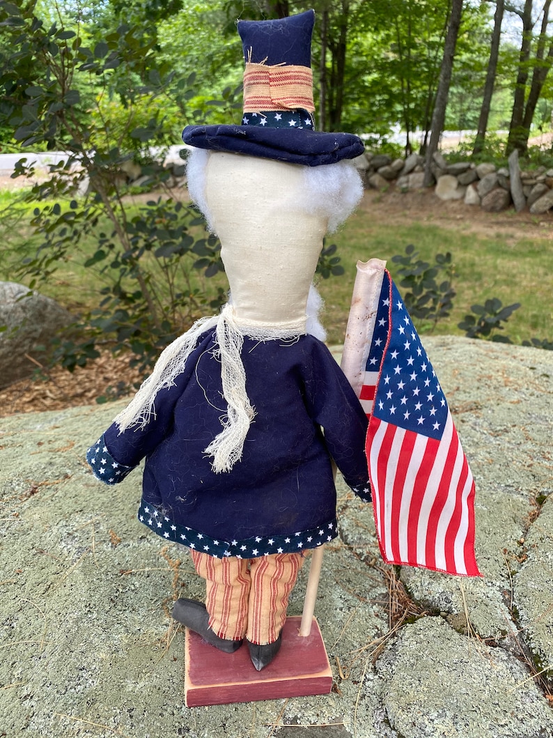 May include: A patriotic Uncle Sam doll, featuring a navy blue coat with white stars, striped pants, and a tall hat. The doll holds a small American flag. The doll is standing on a red wooden base. The doll has white hair and beard.
