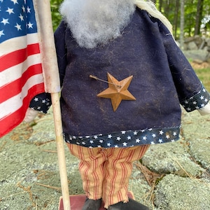 May include: A patriotic doll figure dressed in a navy blue jacket with a bronze star and striped pants. The doll holds a small American flag. The jacket has a white star pattern at the hem. The doll is standing on a red base.