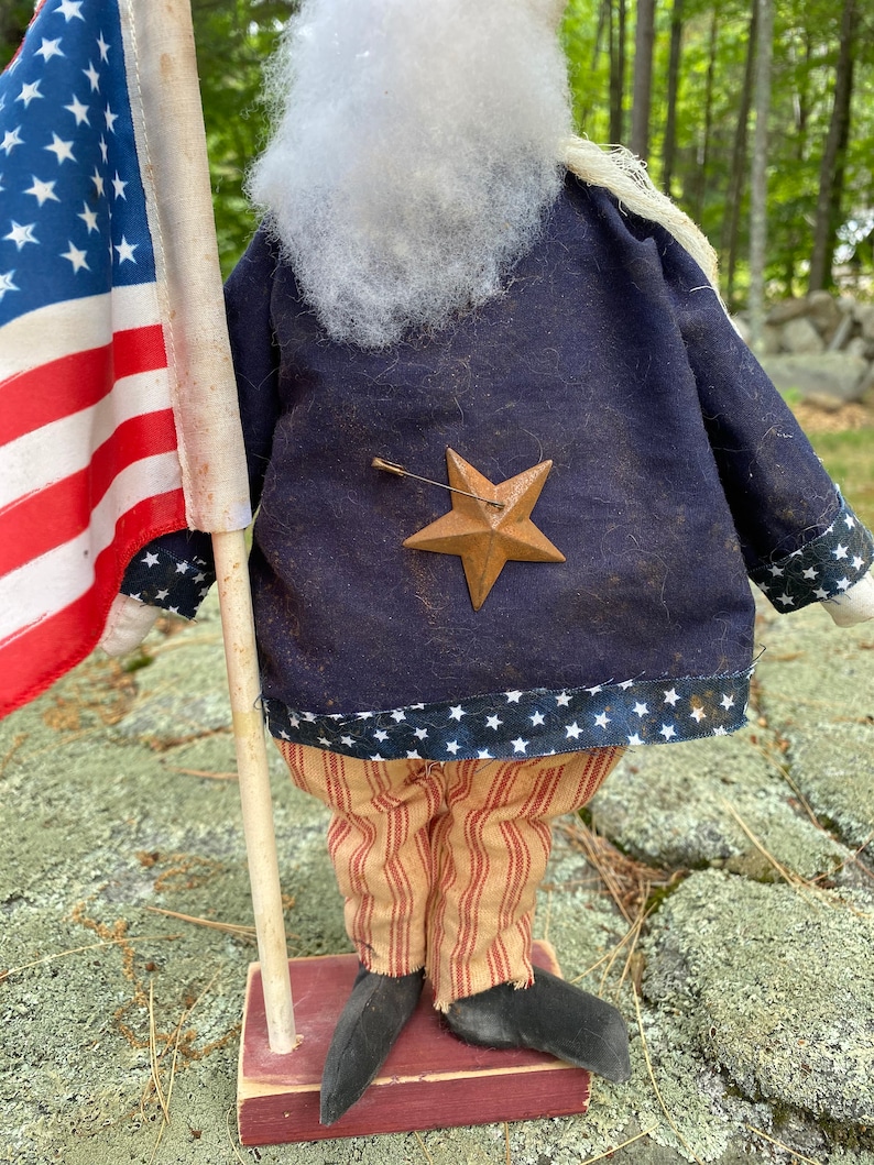 May include: A patriotic doll figure dressed in a navy blue jacket with a bronze star and striped pants. The doll holds a small American flag. The jacket has a white star pattern at the hem. The doll is standing on a red base.