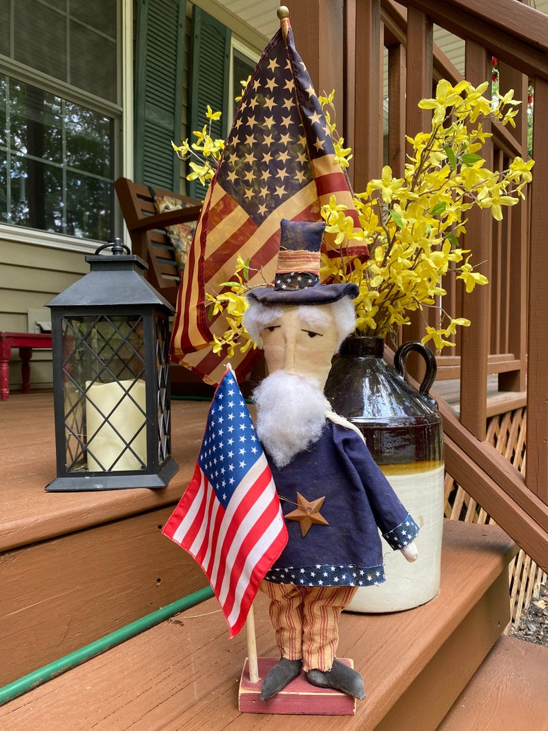 May include: A patriotic Uncle Sam doll stands on a porch, holding a small American flag. He wears a blue coat, striped pants, and a star-adorned hat. An American flag and yellow flowers are in the background.