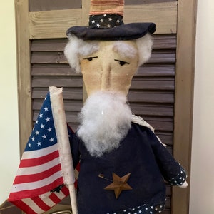 May include: A patriotic Uncle Sam doll with a tall black hat adorned with white stars and a red and white striped band. The doll wears a navy blue coat with white stars and a wooden star pin. It holds a small American flag.