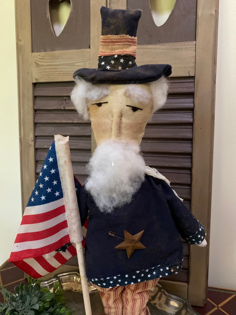 May include: A patriotic Uncle Sam doll with a tall black hat adorned with white stars and a red and white striped band. The doll wears a navy blue coat with white stars and a wooden star pin. It holds a small American flag.