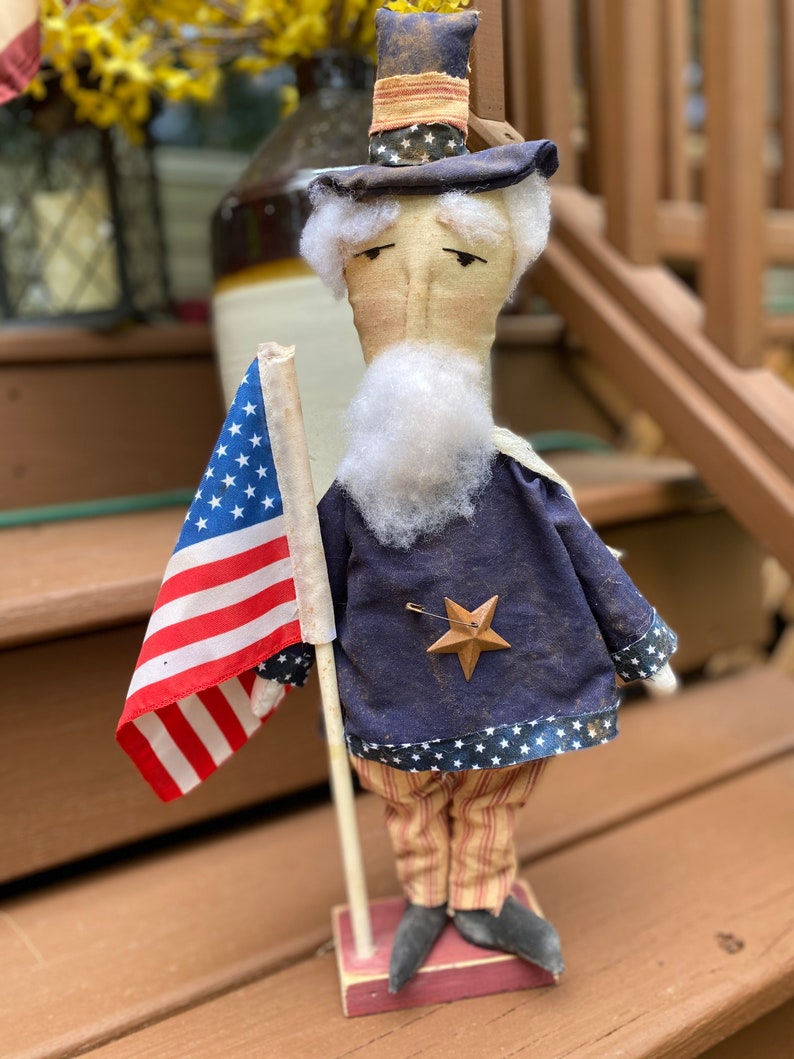 May include: A handcrafted Uncle Sam doll, dressed in a blue coat with a star, striped pants, and a top hat. He holds a small American flag. The doll is standing on a wooden base, perfect for patriotic decor.
