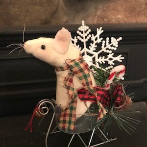 May include: A white plush mouse figurine wearing a plaid scarf, seated in a metal sleigh. The sleigh is decorated with greenery, a candy cane, and a white snowflake. The background is a dark surface and a stone fireplace.