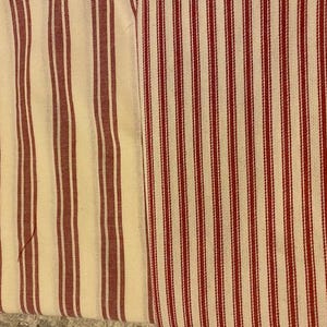 May include: Close-up of two fabric swatches with vertical stripes. One swatch features wide, burgundy stripes on a cream background. The other has narrower, red stripes on a cream background. The fabrics appear to be cotton or linen, suitable for home decor.