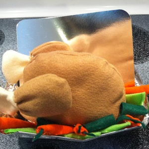 May include: A plush, brown turkey on a foil tray, complete with felt white wings and drumsticks. The tray is garnished with orange carrot and green vegetable accents, creating a festive, handmade Thanksgiving decoration.