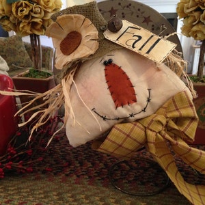 May include: A handcrafted scarecrow decoration with a burlap hat and sunflower accent. The scarecrow has a stitched orange nose and a smiling face. A tag reads "Fall". A red candle and decorative plants are in the background.