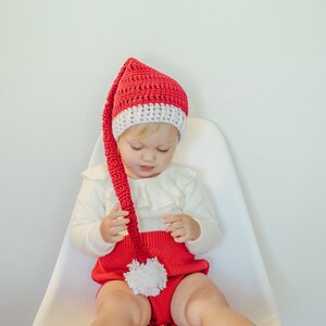 May include: A child wearing a red and white crochet Santa hat with a long, pointed tip. The child is also wearing a red romper with a white pom-pom and a white long-sleeved shirt. The child is sitting on a white chair.