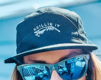 May include: A person wearing a black cap with the text "KRILLIN IT" and a shrimp graphic, and sunglasses with blue reflective lenses. The person has long blonde hair and is wearing a black shirt.