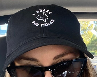 May include: A black baseball cap with white embroidery that reads "I Brake For Molas". The cap is worn by a person with blonde hair and sunglasses.