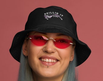 May include: A person wearing a black bucket hat with the text "KILLIN IT" embroidered on the front. The hat has a white brim and a pink flower design. The person is also wearing pink heart-shaped sunglasses and a white long-sleeved shirt.