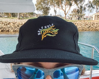 May include: A person wearing a black baseball cap with the text "Help Kelp" and a green kelp design embroidered on the front. The person is also wearing blue sunglasses.