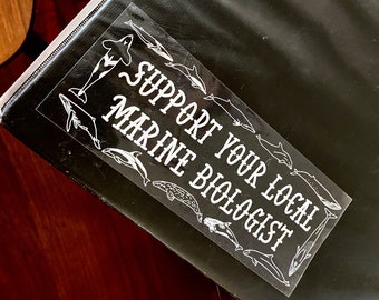 May include: A white sticker on a black background with the text "Support Your Local Marine Biologist" and a variety of white silhouettes of marine animals, including whales, dolphins, and fish.