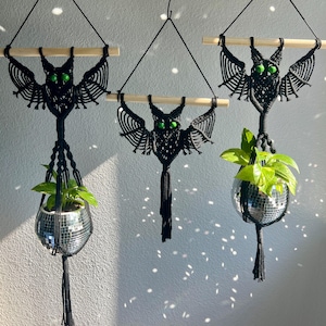 May include: Three black macrame bat plant hangers with green eyes and wooden dowels. Each hanger has a silver disco ball planter with a green plant inside.