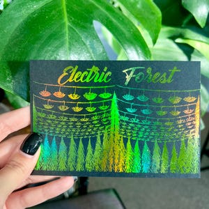 May include: A black card with a holographic design of a forest with trees and string lights that reads "Electric Forest" in gold lettering.