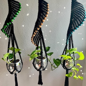 May include: Three black macrame plant hangers with wooden beads and silver disco ball pots. The hangers are hanging from the ceiling and each has a different color of beads: green, brown, and blue.