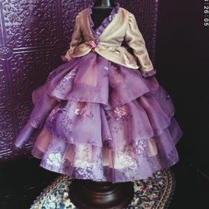May include: A doll dress featuring a layered, tiered purple skirt with a floral pattern and a light beige velvet jacket with long sleeves. The bodice includes a small pink accent at the waist, and the garment is displayed on a black mannequin stand.