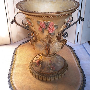 May include: A decorative urn-shaped vase featuring a cream-colored finish with gold accents, adorned with sculpted pink rose floral motifs and green leaves. The vessel includes two dark metal scroll handles and a wide, fluted base with floral details.