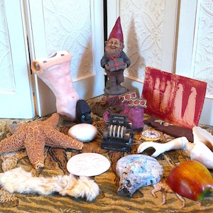 May include: A collection of decorative items including a starfish, a gnome figurine, a seashell, a vintage cash register, and a red and white artwork. The items are arranged on a patterned rug, creating a whimsical display.
