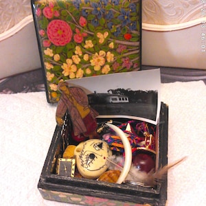 May include: A small, open wooden box with a variety of objects inside, including a small clock, a carved face, a white crescent, and a photo. The box is decorated with floral patterns. A matching box with a floral design is in the background.