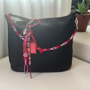 May include: Black handbag with a textured surface and a decorative chain strap in shades of pink and red. The bag features a keychain with the word "AMOR" and a heart charm. A small red tag is also attached.