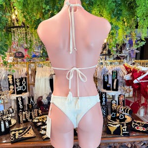 May include: Ivory bikini set on a mannequin. The halter top and low-rise bottoms feature a textured pattern and tassel accents. The background includes jewelry displays and greenery.