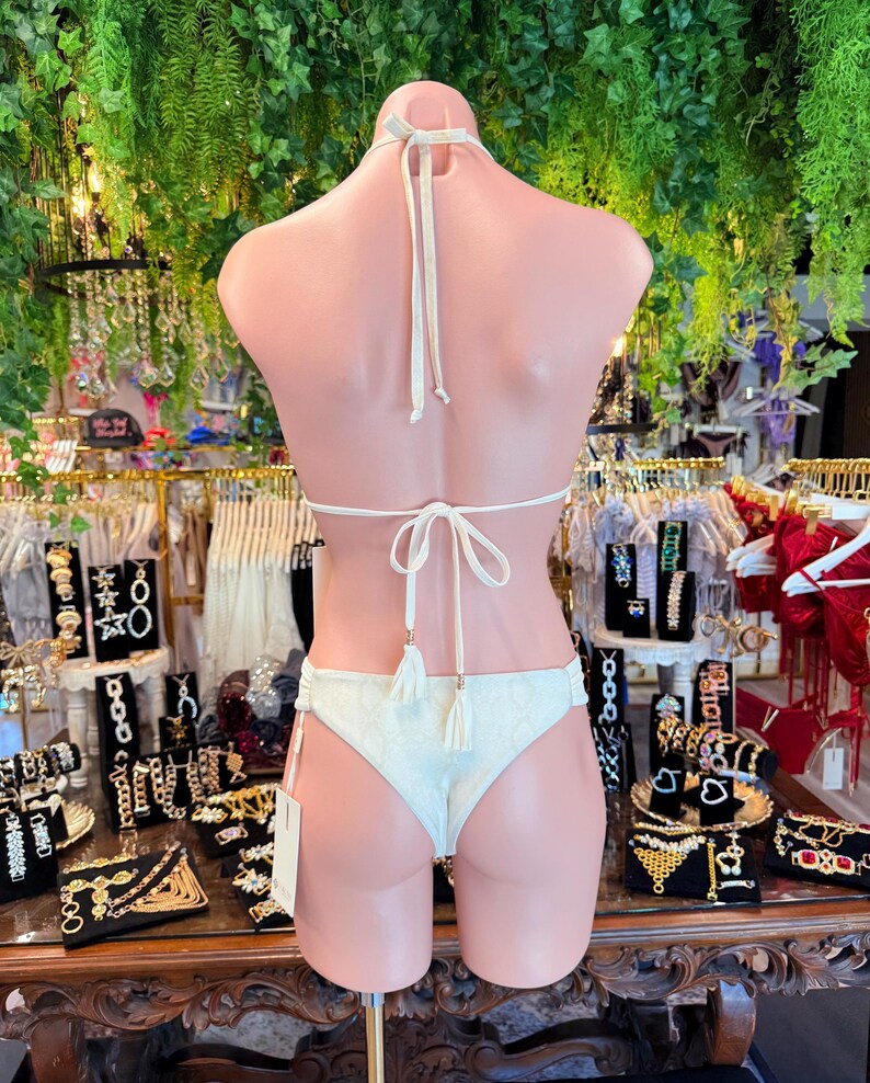 May include: Ivory bikini set on a mannequin. The halter top and low-rise bottoms feature a textured pattern and tassel accents. The background includes jewelry displays and greenery.
