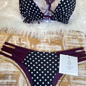 Puede incluir: Un conjunto de bikini de lunares blancos y negros. La parte superior es de estilo triangular con tirantes finos. La parte inferior es de talle bajo con múltiples correas burdeos en los laterales. El conjunto incluye una etiqueta con el nombre de la marca.