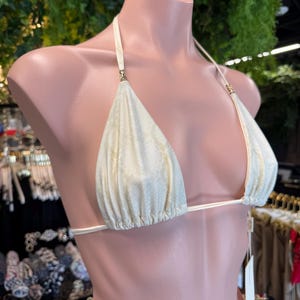 May include: Cream-colored bikini top with a subtle snakeskin pattern. The triangle cups are connected by thin, white straps that tie at the neck and back. Gold-colored accents are visible at the strap attachments. The bikini is displayed on a mannequin.