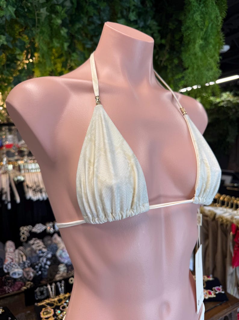 May include: Cream-colored bikini top with a subtle snakeskin pattern. The triangle cups are connected by thin, white straps that tie at the neck and back. Gold-colored accents are visible at the strap attachments. The bikini is displayed on a mannequin.