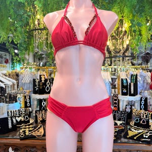 May include: A red bikini set displayed on a mannequin. The halter top features embellished detailing along the neckline. The bottoms are a classic brief style. The background includes jewelry displays and greenery.