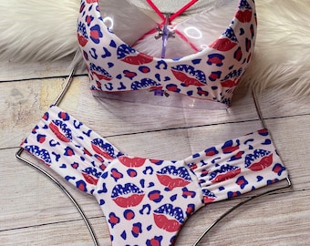May include: White bikini with a red, white, and blue lip print. The bikini top has a criss-cross back strap and the bottom has a high-cut leg.