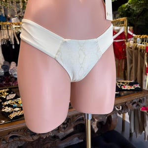 May include: Cream-colored bikini bottom with a snakeskin print center panel. The bikini bottom is displayed on a mannequin. The fabric appears smooth and the design is simple and elegant.