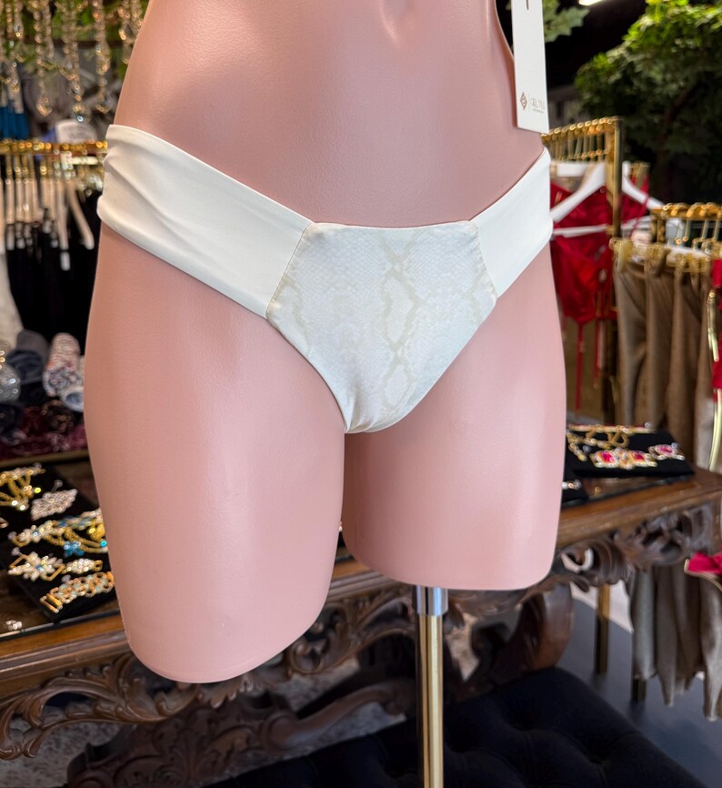 May include: Cream-colored bikini bottom with a snakeskin print center panel. The bikini bottom is displayed on a mannequin. The fabric appears smooth and the design is simple and elegant.