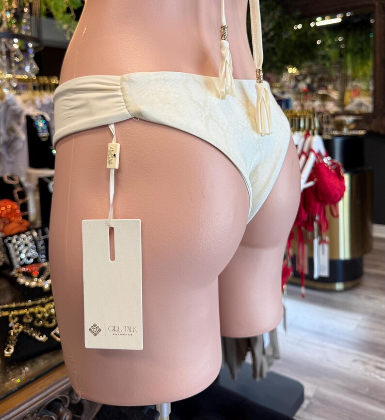May include: Off-white bikini bottom with a textured pattern, featuring a ruched design on the sides. The bikini has cream-colored tassels and a tag with the brand name. The swimwear is displayed on a mannequin.