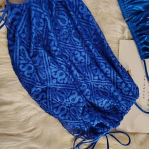 May include: A bright blue lace bikini bottom featuring a geometric pattern and side tie closures. The garment is paired with a matching metallic blue bikini top and a white product tag with visible text, presented as a two-piece swimwear set.