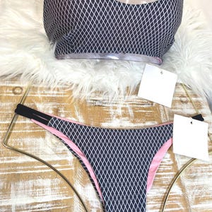 May include: A two-piece swimsuit set featuring a navy blue diamond pattern with white accents. The top has a transparent overlay and black straps. The bottoms are pink-lined. The set is displayed on a wooden surface.