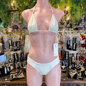 May include: A two-piece bikini set on a mannequin. The top is a triangle style with thin straps and gold-colored accents. The bottom is a low-rise style with a textured panel. The bikini is a light, shimmering color.