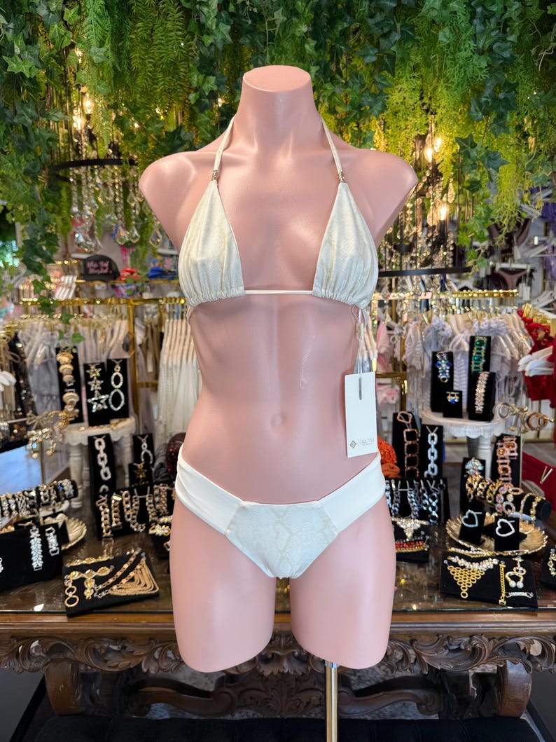 May include: A two-piece bikini set on a mannequin. The top is a triangle style with thin straps and gold-colored accents. The bottom is a low-rise style with a textured panel. The bikini is a light, shimmering color.