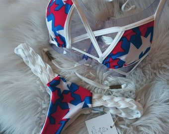 May include: A white bikini top and bottom with red and blue stars. The top has a criss-cross design and the bottom has a braided strap. The tag says "unica swimwear".
