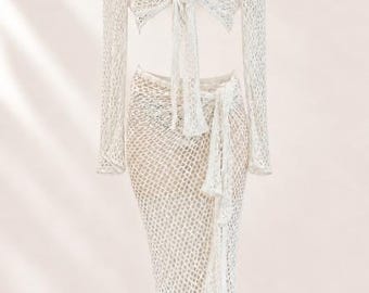 May include: A two-piece set consisting of a long-sleeved, white, open-knit crochet top with a front tie closure and a matching long, white, open-knit crochet skirt featuring a side tie detail. Both pieces have a loose, net-like texture.