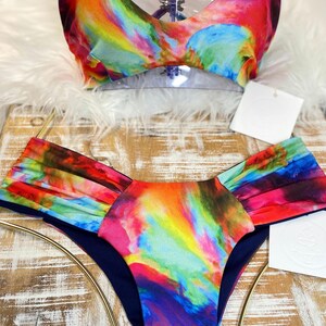 May include: A vibrant two-piece swimsuit set. The top is a V-neck style with thin straps. The bottoms are a high-waisted design. Both pieces feature a colorful, abstract pattern with shades of red, blue, yellow, and purple.