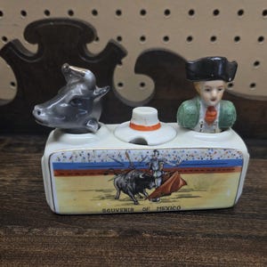 Puede incluir: Un recuerdo de cerámica de México con un toro, un matador y una cabeza de toro. La pieza rectangular tiene una base blanca con una escena de corrida pintada y las palabras "Souvenir of Mexico". Un sombrero pequeño y una figura de matador están en la parte superior.