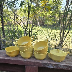 May include: A collection of yellow vintage dishware, including soup bowls and a covered serving dish, arranged on a wooden surface. The bowls are stacked and have handles. The scene is set outdoors with a natural background of trees and foliage.