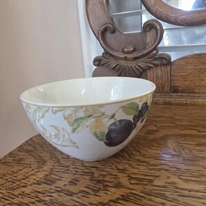 May include: A white ceramic bowl with a fruit design featuring plums and leaves. The bowl has a wide rim and a slightly flared shape. The fruit design includes purple plums, green leaves, and gold accents. The bowl is on a wooden surface.