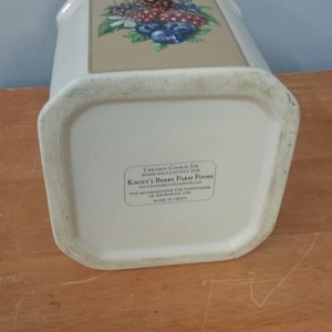K&ouml;nnte beinhalten: Ein wei&szlig;er Keramik-Keksbeh&auml;lter mit rechteckiger Form und einem Blumendesign mit Beeren und Blumen. Das Gef&auml;&szlig; hat ein Etikett mit Text auf der Unterseite. Das Gef&auml;&szlig; steht auf einer Holzoberfl&auml;che.