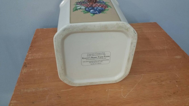 K&ouml;nnte beinhalten: Ein wei&szlig;er Keramik-Keksbeh&auml;lter mit rechteckiger Form und einem Blumendesign mit Beeren und Blumen. Das Gef&auml;&szlig; hat ein Etikett mit Text auf der Unterseite. Das Gef&auml;&szlig; steht auf einer Holzoberfl&auml;che.
