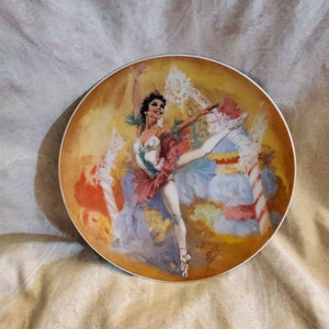 May include: A porcelain plate featuring a colourful painting of a ballerina in a white tutu and pink and green skirt. The ballerina is dancing in a swirl of colours and lights. The plate has a gold rim and a signature in the bottom right corner.